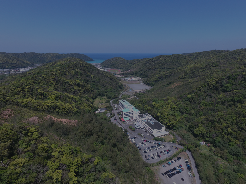 海と山に囲まれた施設の空撮。駐車場と建物が見える。